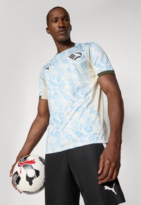 Uomo che tiene in mano un pallone da calcio Puma bianco e nero, indossando una maglia a maniche corte azzurro chiaro con motivo paisley e pantaloni neri Puma, su uno sfondo semplice.