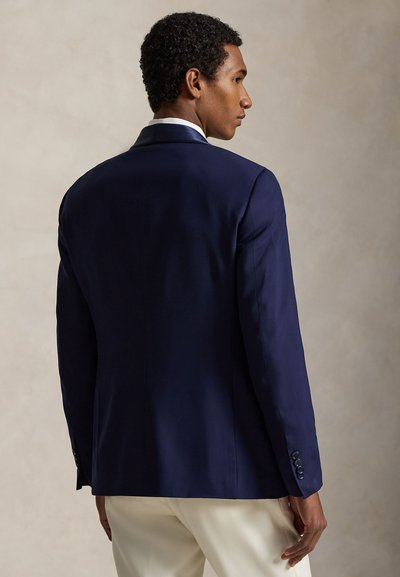 Férfi, aki tengerészkék blézert és fehér nadrágot visel, hátrafelé nézve, egyszínű bézs háttér előtt.