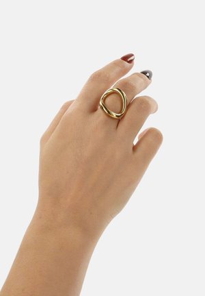 Goudkleurige metalen ring met een vloeiende, organische vorm en een hol midden. Draagt aan een hand met gepolijste nagels, met een gladde afwerking als uitstraling.