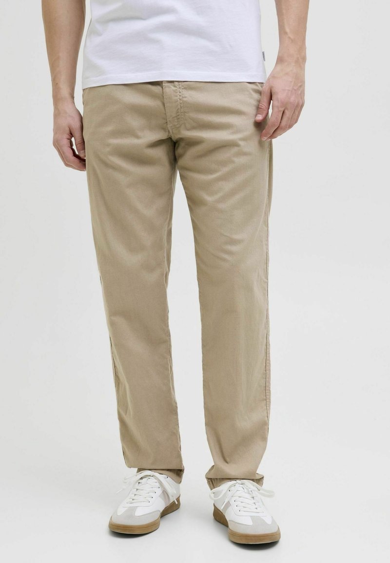 Beiget puuvillaiset housut, joissa on rento istuvuus, sivutaskut ja vähäiset saumaustiedot, yhdistettynä valkoisiin tennareihin, joissa on vaaleanruskeita yksityiskohtia.