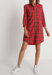 Robe chemise à carreaux rouges avec fermeture à boutons, dotée de manches trois-quarts et d'une coupe droite, associée à des baskets blanches.