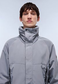 Light grey high-collar jacket with a smooth texture, side zippers, and contrasting stitching detail. Features a sporty, modern design.