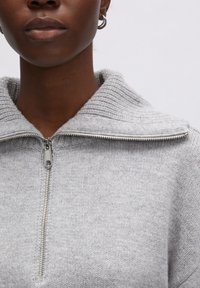 Grijze, gestructureerde trui met een brede, ribbelkraag en een voorkant met ritssluiting. Heeft een zachte gebreide structuur die de nadruk legt op warmte en comfort.