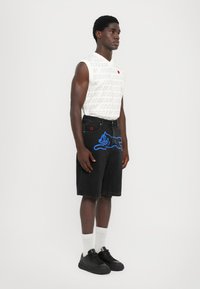 Camiseta sin mangas blanca con el texto "HELADO" repetido, combinada con shorts de mezclilla negros que presentan un gráfico de un gato azul. Zapatillas negras y calcetines blancos.