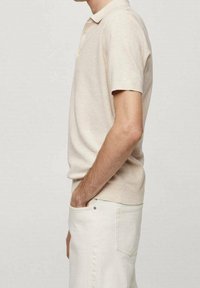 Beżowa koszulka polo z teksturowanym wzorem, krótkimi rękawami i kołnierzykiem. Noszona z jasnymi spodniami, ukazująca profil boczny.