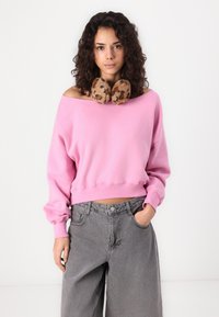 Felpa cropped rosa con design a spalla aperta, abbinata a jeans larghi grigi. La modella indossa un scaldacollo in pelliccia maculata marrone.