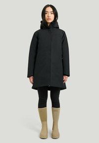 Svart vattentät kappa med huva, med rak skärning, långa ärmar och framzip. Bärs över svarta leggings och beige regnstövlar.