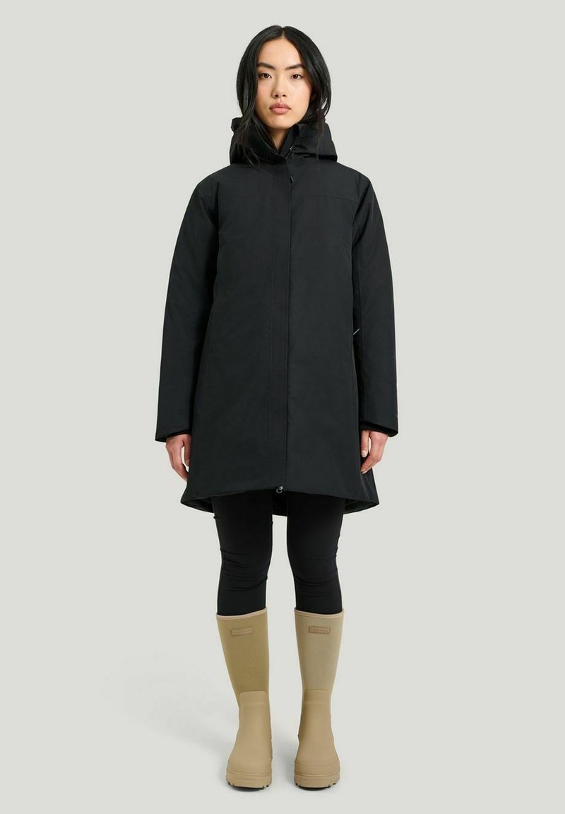 Svart vattentät kappa med huva, med rak skärning, långa ärmar och framzip. Bärs över svarta leggings och beige regnstövlar.
