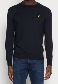 Mann in einem enganliegenden schwarzen Langarmpullover mit einem kleinen gelben Vogel-Logo auf der Brust, kombiniert mit dunkelblauen Jeans.