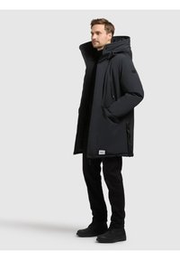 Palton puffer lung, negru, cu o glugă mare, buzunare cu fermoar și o textură netedă. Prezintă un patch cu logo deasupra buzunarului stâng.