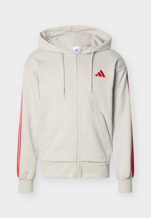 Beige zip-up hoodie with front pockets, red Adidas logo on chest, and three red stripes down each sleeve. Drawstring hood included.