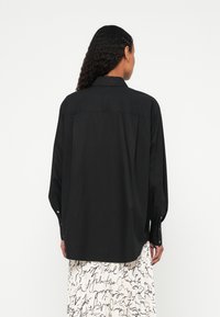 Γυναίκα με μακριά σγουρά μαλλιά φοράει χαλαρό μαύρο πουκάμισο με κουμπιά και λευκή πλισέ φούστα με μαύρα σχέδια γραφής, με την πλάτη προς τα εμάς.