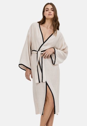 Femme portant une robe beige avec une bordure noire, liée à la taille, avec des manches longues et une fente mi-cuisse, debout devant un fond uni.