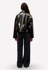 Vrouw met lang bruin haar staat met haar rug naar je gekeerd, draagt een kort zwart leren jack en hoge, wijde zwarte jeans.