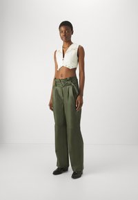Crop top bianco con zip anteriore, hardware argentato e cuciture sottili. Pantaloni beige a vita alta, larghi, con dettaglio della cintura.