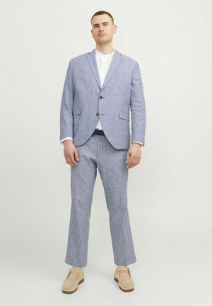 Completo in lino blu chiaro con giacca monopetto e pantaloni a gamba dritta, abbinato a una camicia bianca e scarpe beige.