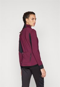 Veste de sport bordeaux avec un col haut, des panneaux latéraux noirs et une poche zippée. Tissu texturé et design ajusté pour le confort.