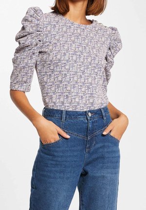 Femme portant un haut texturé à manches courtes bouffantes, rentrée dans un jean bleu taille haute, les mains dans les poches.