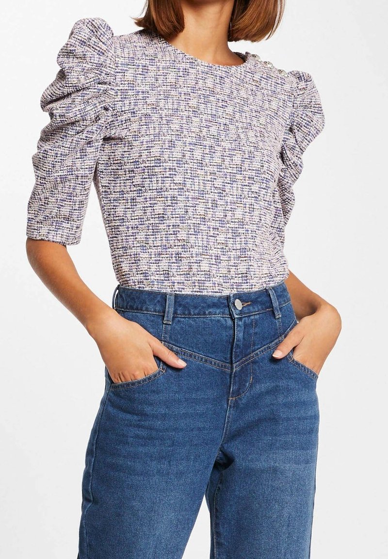 Femme portant un haut texturé à manches courtes bouffantes, rentrée dans un jean bleu taille haute, les mains dans les poches.