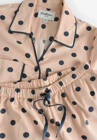 Conjunto de pijama en tejido melocotón con lunares azules marinos. Presenta una parte superior con cuello, un bolsillo y pantalones cortos con cintura elástica y cordón de ajuste.