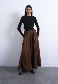 Γυναίκα με μακριά σκουρόχρωμα μαλλιά, που φοράει μαύρο μπλουζάκι με μακριά μανίκια, καφέ φούστα ψηλόμεση και μαύρες μυτερές μπότες, που στέκεται μπροστά σε λευκό τοίχο.