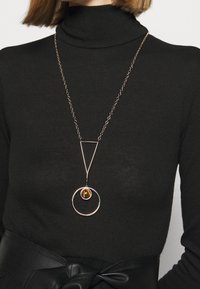 Collier doré avec un pendentif circulaire et une perle d'ambre, suspendu à une chaîne, porté sur un col roulé noir.
