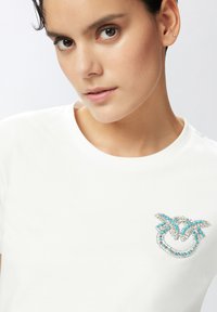 Camiseta de algodón blanca con un intrincado diseño de rhinestones azules y plateados en el lado izquierdo del pecho, escote redondo y mangas cortas.