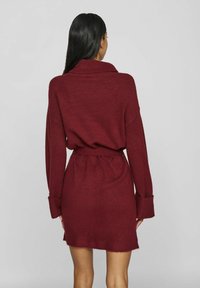 Robe en maille bordeaux à manches longues, col châle et taille ceinturée ; présente des poignets côtelés et une texture duveteuse.