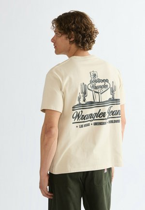 Uomo con capelli ricci che indossa una maglietta beige Wrangler Jeans con stampa di un cactus e le scritte Las Vegas, Greensboro, Worldwide sulla schiena, rivolto verso sinistra.