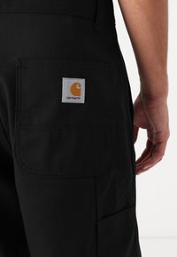 Schwarze Arbeitshose mit einer Gesäßtasche, die mit einem grauen quadratischen Aufnäher mit einem orangefarbenen Logo versehen ist, aus strapazierfähigem Stoff gefertigt.