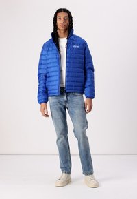 Blauer Puffermantel mit horizontalem Steppmuster, hohem Kragen, Frontreißverschluss, kombiniert mit hellblauen Jeans und cremefarbenen Sneakers.