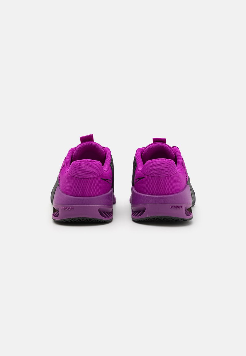 Zapatillas deportivas negras y moradas con un acabado texturizado, cuello acolchado y una pestaña para tirar. Cuenta con una suela prominente y un logo en el talón.