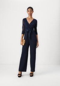 Mono de encaje y tejido azul marino con escote en V profundo, mangas tres cuartos y cintura atada; combinado con un clutch dorado y tacones negros.