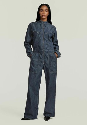 Vrouw met lang gevlochten haar, gekleed in een donker denim jumpsuit met rits aan de voorkant en handen in de zakken, staand op een neutrale achtergrond.