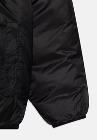 Chaqueta de plumas negra con un exterior liso y brillante, puños elásticos y un bolsillo lateral. El material parece ligero y aislante.