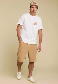 T-shirt en coton blanc avec un graphique rouge sur la poitrine gauche, associé à un short en velours côtelé beige et des chaussures blanches. L'arrière-plan est beige.