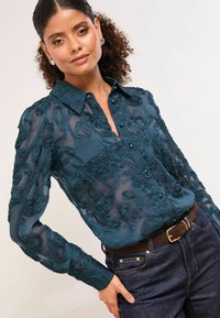 Mäkká tyrkysová blúzka s kvetinovou výšivkou, gombíkovým zapínaním a golierom. Dlhé štruktúrované rukávy v kombinácii s tmavomodrými džínsami a hnedým opaskom.