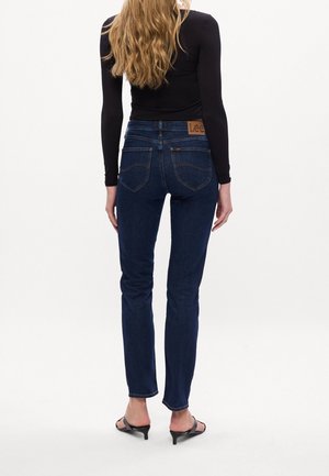 Frau mit langen blonden Haaren, die dunkelblaue Jeans, ein schwarzes langärmliges Oberteil und schwarze Sandalen mit Absatz trägt, steht mit dem Rücken zur Kamera vor weißem Hintergrund.