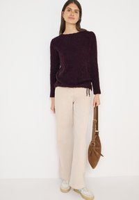 Bordowy sweter z teksturą i długimi rękawami, beżowe szerokie spodnie oraz brązowa zamszowa torba. Outfit uzupełniają sneakersy z jasnoniebieskimi akcentami.