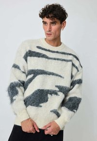 Pull-over en maille douce crème et grise avec des motifs abstraits ondulés. Col rond, poignets et ourlet côtelés. Fini texturé.