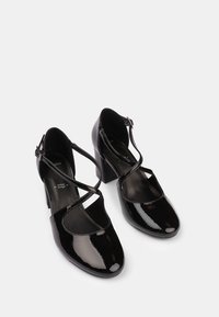 Chaussures à talons en cuir verni noir avec un bout arrondi, un design à brides croisées et une boucle à la cheville. Texture lisse et talon carré pour plus de stabilité.