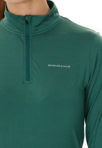 Grøn langærmet træningsbluse lavet af glat, letvægtsstof; har en lynlåset krave og "ENDURANCE" logoet på fronten.