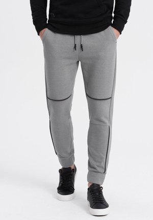 Mann trägt graue Jogginghosen mit gerippten Kniepaneelen, schwarze Sneakers und einen schwarzen Hoodie, steht mit den Händen in den Taschen.