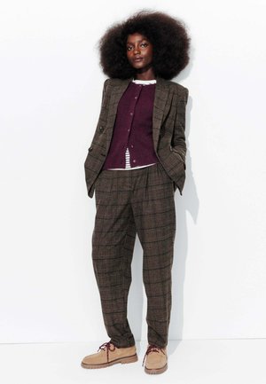 Costume à carreaux marron avec une veste ajustée et un pantalon. Superposé avec un cardigan en maille bordeaux sur une chemise rayée blanche. Chaussures beiges. Cheveux bouclés.