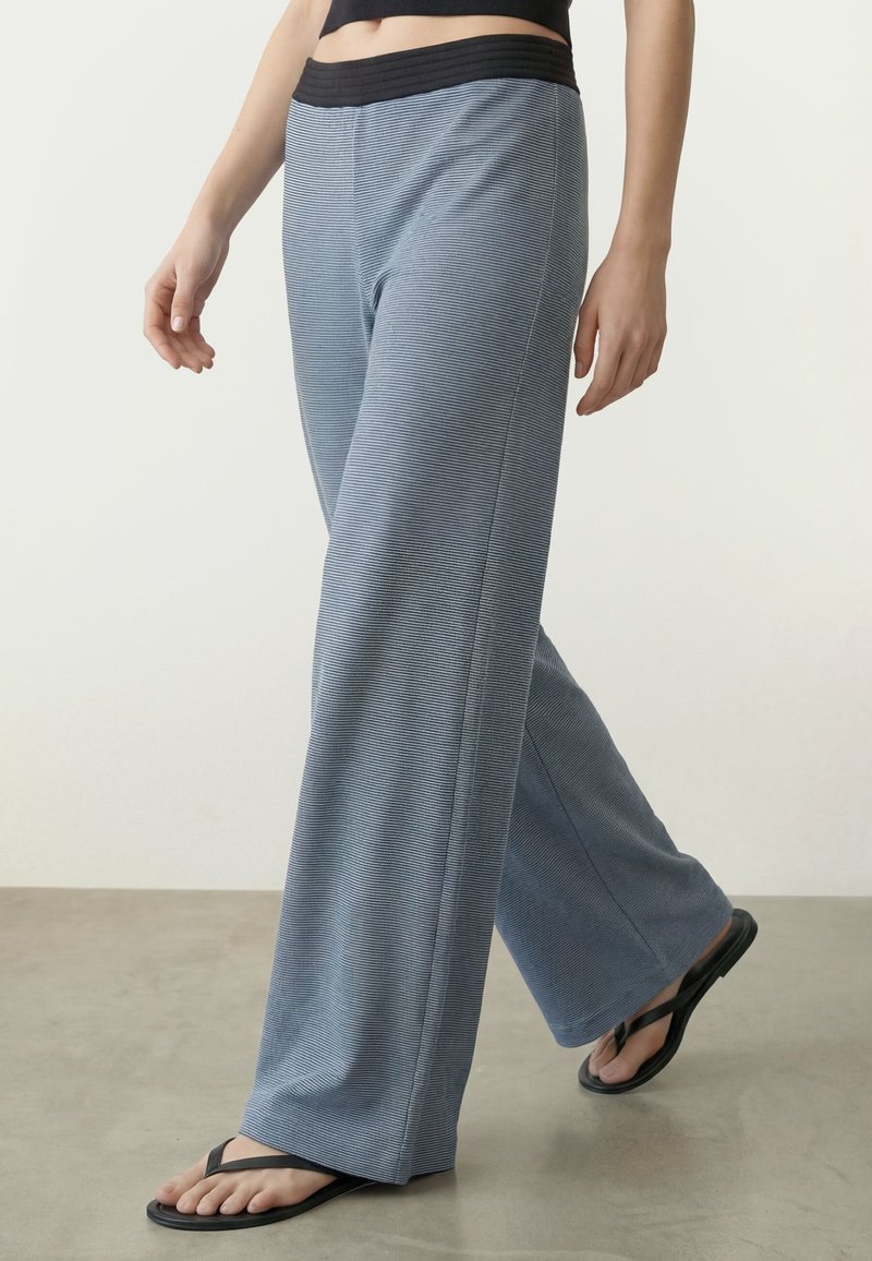 Partie inférieure du corps d'une personne marchant, portant un pantalon bleu rayé ample avec une ceinture noire et des tongs noires sur un sol gris.