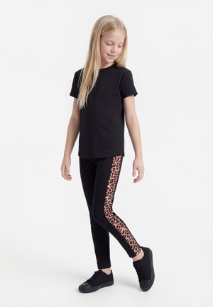 Fille posant, portant un t-shirt noir et des leggings noirs avec des bandes à motif léopard sur les côtés, assortis à des baskets noires.
