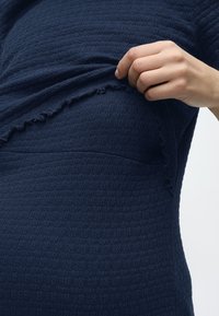 Ruka zdvíhajúca tmavomodrú textúrovanú látku priliehavého odevu, odhaľujúca jemný nariasený okraj a zodpovedajúci pás pod ňou.