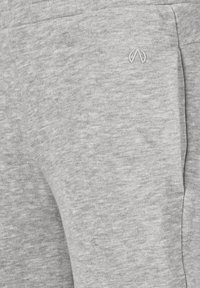 Nærbillede af en lys grå stoflomme med et lille, sølvfarvet logoemblembroderet på den øverste højre side.