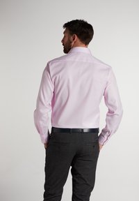 Helles rosa Hemd mit langen Ärmeln und Knopfmanschetten, kombiniert mit dunkelgrauen gemusterten Hosen und einem schwarzen Gürtel, von hinten betrachtet.