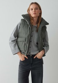 Grüne gesteppte Weste mit einem kuscheligen, fellgefütterten Kragen über einem grauen gerippten Pullover. Das Model trägt dunkle Jeans, die einen lässigen Schichtlook zeigen.